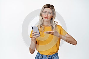 Troubled young woman asking help with mobile phone, pointing at smartphone and looking upset, standing over white