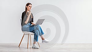 Troubled young woman sitting on chair with laptop, making mistake in online project, thinking over solution to problem