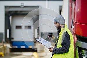 Truck driver standing by red truck and holding clipboard, looking at cargo details, delivery schedule.