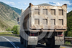 Truck with a gray trailer with rubble in the back of a dangerous highway against the background of rocky mountains and blue sky.