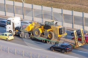 Truck with a long trailer platform for transporting heavy machinery, loaded tractor with a bucket. Highway transportation traffic