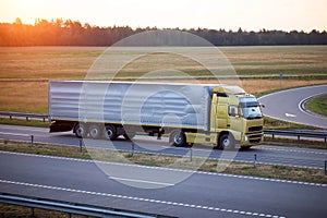 A truck with a tilt semi-trailer carries cargo along the highway in the evening against the backdrop of a summer sunset