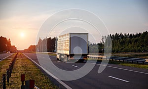 A truck with a tilt semi-trailer carries cargo along the highway in the evening against the backdrop of a summer sunset