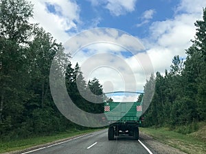 A truck, a tractor with a large green trailer is driving along a forest asphalt road with green trees on the grounds