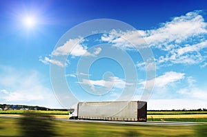 Truck with the trailer on the countryside road in motion with fields and trees against blue sky with clouds and sun