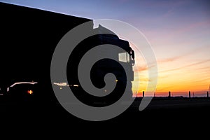A truck with a trailer rides on the motorway at night against the backdrop of an orange sunny sunset. The concept of the