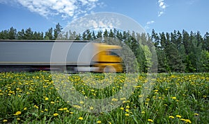 A truck with a yellow cab rushes along the road along the forest, the roadside is covered with yellow dandelions