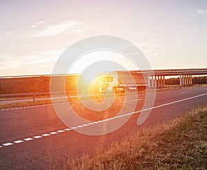 A trucker driver transports cargo on a white wagon against the background of a road bridge and a sunset. Shipping, business and