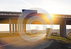 Trucks with semi-trailers move along the highway against the backdrop of a sunny sunset. Automobile tie and bridge on the