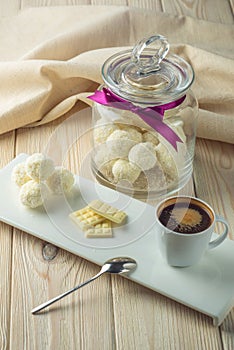 Truffle balls with white chocolate sprinkled with coconut on a saucer next to a jar of candy and a Cup of coffee