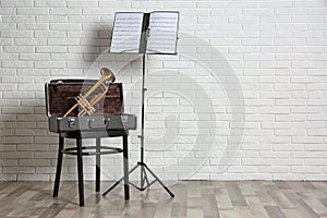 Trumpet, chair, case and note stand with music sheets near brick wall.