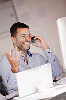 Trust him to cut the deal. an expressive young man sitting at his desk and talking on his mobile phone.