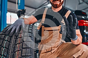 Trustworthy mechanic showing ok gesture, holding car tire and smiling on the car repair shop