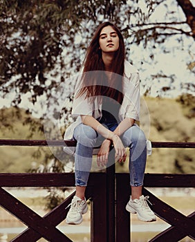 Tthinking teen casual woman in blue jeans sitting on the fence outdoors on summer green trees background. Closeup portrait in