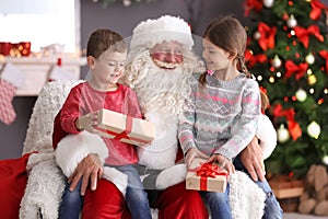 Little children with gift boxes sitting on Santa Claus` knees indoors