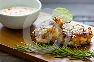TTwo shrimp cakes on a cutting board with a lemon garlic dipping sauces lemon wedge and a stem or rosemary on a wooden table..