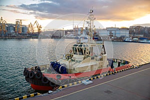 A tug of a ship stands moored at the pier on a summer evening during sunset. Sea harbor. Port cranes unload cargo