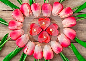 Tulip flowers are laid out in the shape of a heart on a wooden background, among them are lit candles in the shape of a heart.