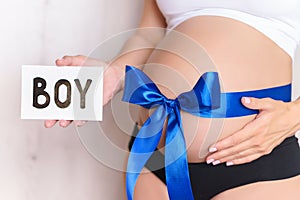 Tummy with blue ribbon and bow of pregnant caucasian woman which is holding nameplate boy