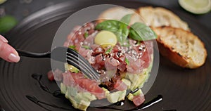 Tuna and avocado tartare with sesame seeds, capers and egg yolk on a dark ceramic plate with baguette bread crouton