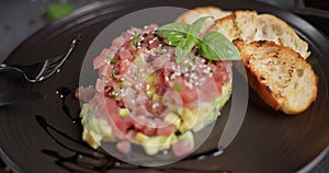 Tuna and avocado tartare with sesame seeds, capers and egg yolk on a dark ceramic plate with baguette bread crouton
