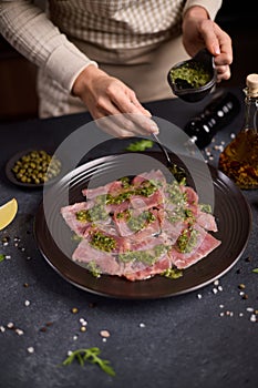 tuna carpaccio - woman puts capers green sauce onto slices of fresh raw tuna fillet on black ceramic plate