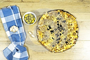 tuna pizza with cheese and tomato and olives on cutting wooden board with green olive bowl and garlic.