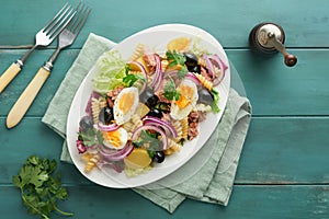 Tuna salad with pasta eggs potatoes olives red onions sauce in white plate on old turquoise wooden rustic table background Nicoise