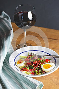 Tuna salad with vegetables, boiled eggs, cherry tomatoes and spices and one glass of red wine over wooden background