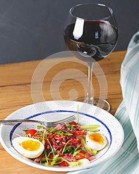 Tuna salad with vegetables, boiled eggs, cherry tomatoes and spices and one glass of red wine over wooden background