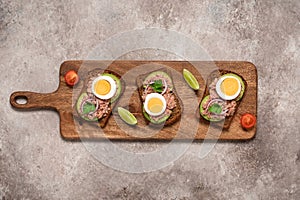 Tuna toast. Open sandwich with tuna, rye bread, avocado and boiled egg on a wooden long board, brown grunge background. Top view,