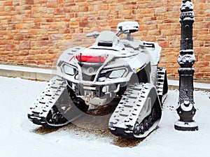 A tuned tracked ATV covered with snow stands near a stone wall