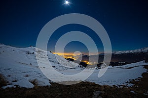 Turin city lights, night view from snow covered Alps by moonlight. Moon and Orion constellation, clear sky. Italy.
