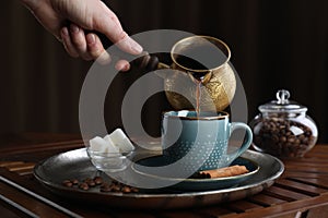 Turkish coffee. Woman pouring brewed beverage from cezve into cup at wooden table, closeup