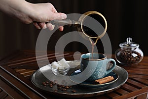 Turkish coffee. Woman pouring brewed beverage from cezve into cup at wooden table, closeup