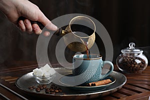 Turkish coffee. Woman pouring brewed beverage from cezve into cup at wooden table, closeup