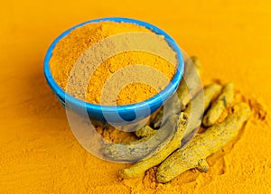 Turmeric powder in blue bowl with dry roots at orange background