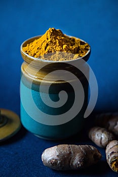 Turmeric Powder in a Ceramic Jar surrounded by Haldi Roots on a Blue Background. Yellow Southeast Asian Spice.