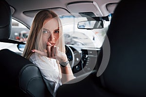 Turned back to the cameramen at backseat. Beautiful blonde girl sitting in the new car with modern black interior