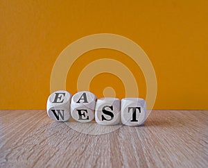 Turned wooden cubes and changes black word east to west, or vice versa. Beautiful wooden table, orange background, copy space.