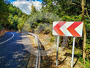 Turning traffic sign for incoming curves with metal fence barrier on the side of the new asphalt road surrounded by green trees