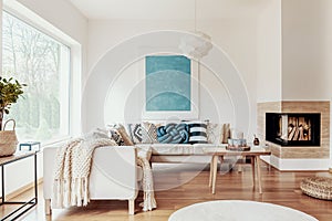 Turquoise blue knot pillow on a beige corner sofa and an abstract poster on a white wall in a modern living room interior