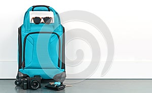 Turquoise blue travel luggage with sunglasses, camera, wallet, and keys against a white wall. Ready for a vacation holiday.