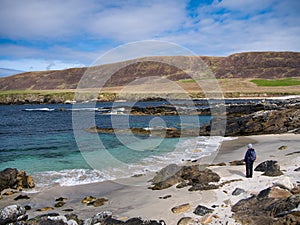 With turquoise, clear water and white sand, a single female walker looks to the sea on a beach at Collaster, Shetland, UK