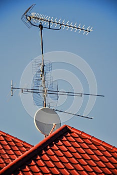 TV antennas and satellite dish for television mounted on the tiled roof of house isolated on blue sky background in countryside