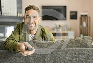 TV concept of a Handsome cheerful young man holding remote control and watching TV while sitting on the couch at home