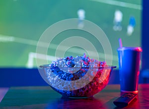 TV remote control, popcorn in a bowl and a drink in a glass on the background of a football match on TV. Neon lighting.