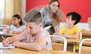 Tween boy listening to schoolteacher and writing in notebook in class