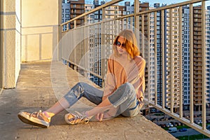 Tween redhead girl in pullover, jeans and sneakers sitting on balcony against high-rise multi-storey residential building at