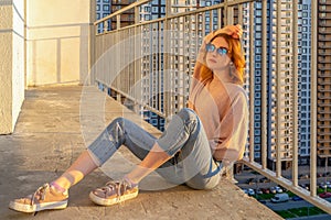 Tween redhead girl in pullover, jeans and sneakers sitting on balcony against high-rise multi-storey residential building at